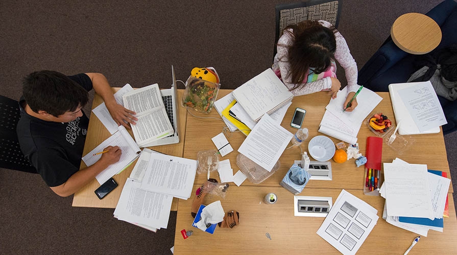 Two students study at a table surrounded by papers and notebooks
