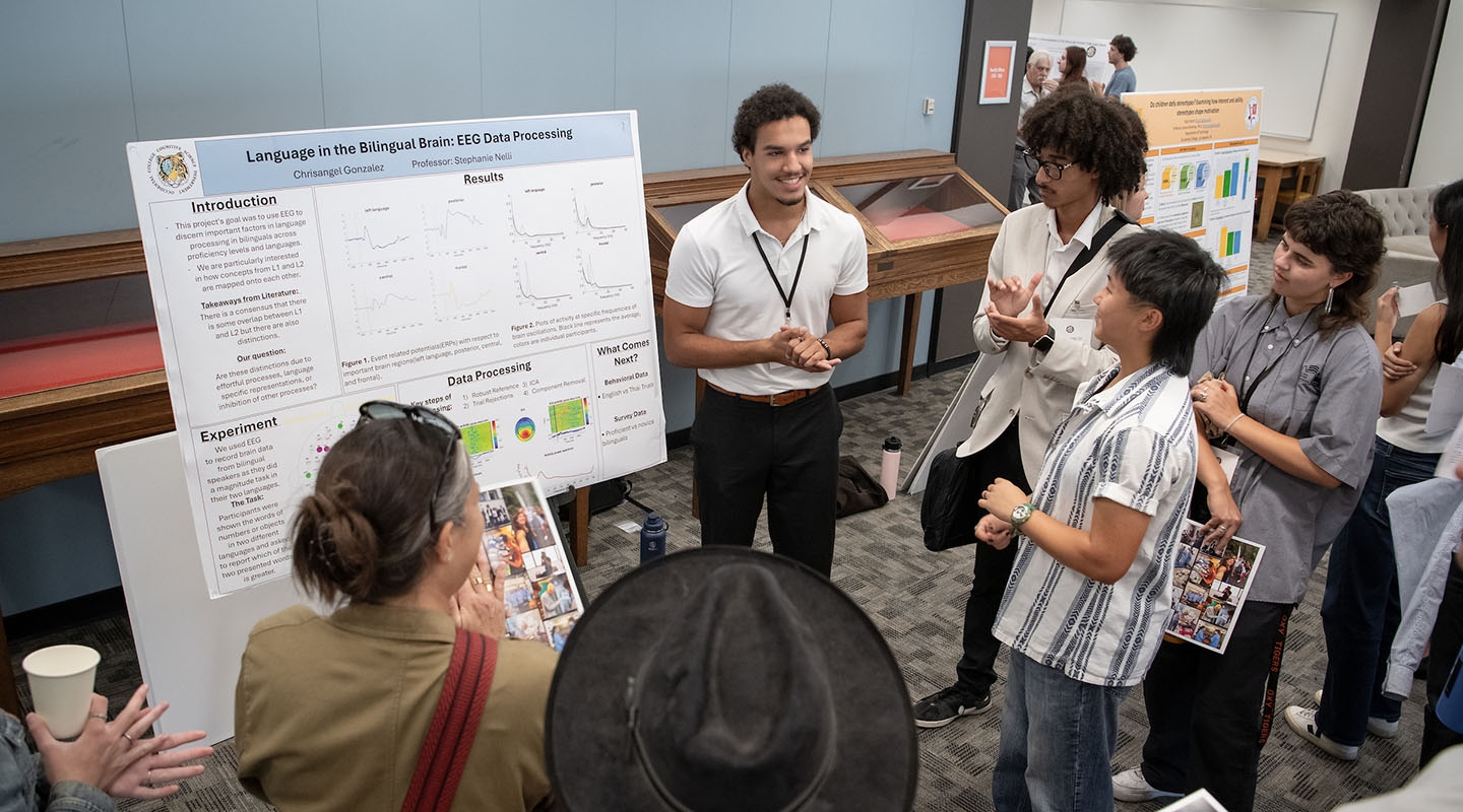a group of Occidental College students stands around a male student giving a poster presentation about his research