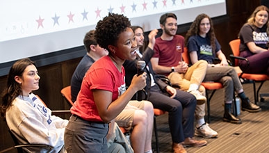 a student stands with a mic in front of a panel of other students