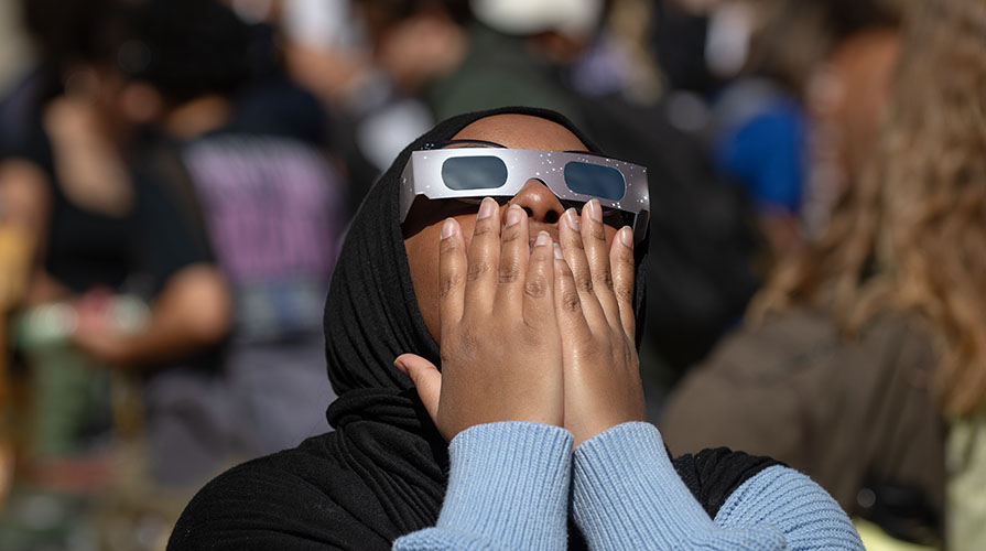 a young woman wearing solar eclipse glasses holds her hands to her mouth in surprise as she looks up at the eclipse in the sky