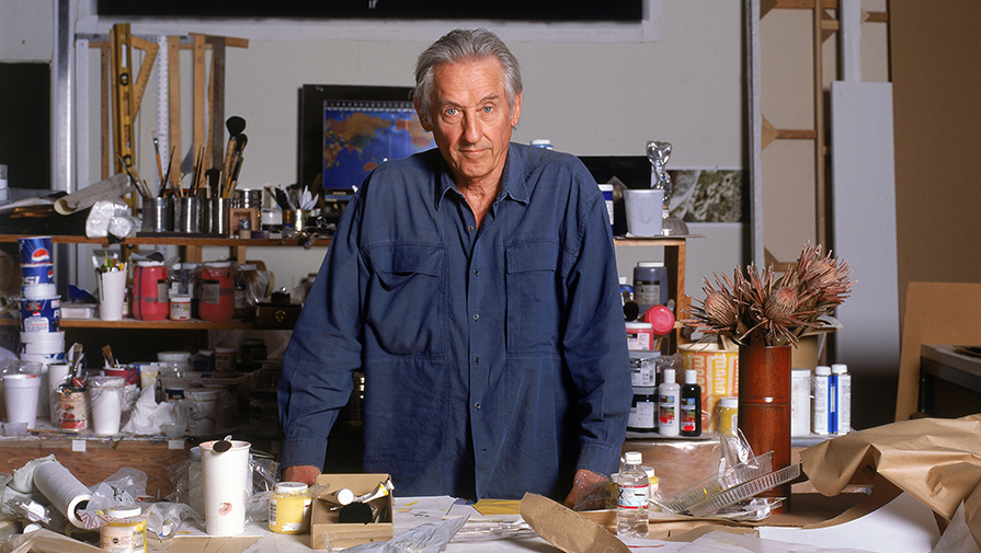 Ed Ruscha in his art studio with supplies, wearing a blue smock.