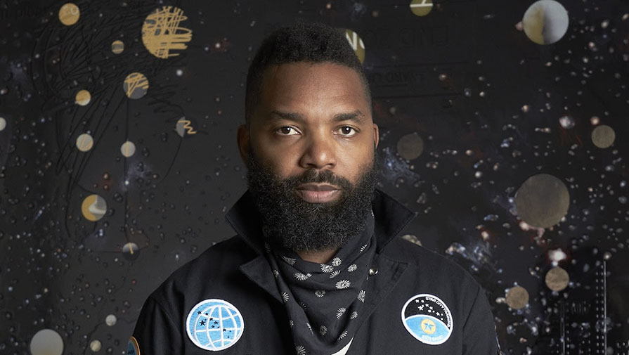 Tavares Strachan in a black suit with cosmic imagery behind him on a black background