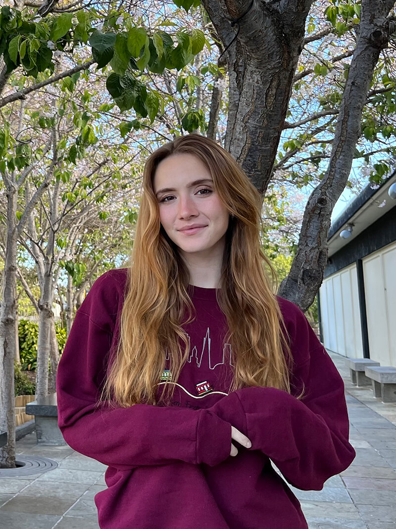 Frannie is wearing a Burgundy sweatshirt and is standing on a walkway with several trees in the background