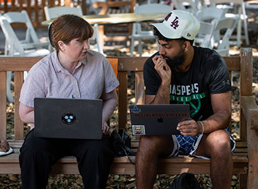 An Occidental College professor sits next to a student as they look at their laptops together