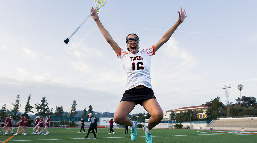 An Occidental College lacrosse player jumps into the air triumphantly, smiling.