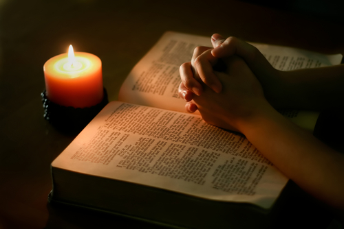 Two hands folded over a Bible illuminated by candlelight