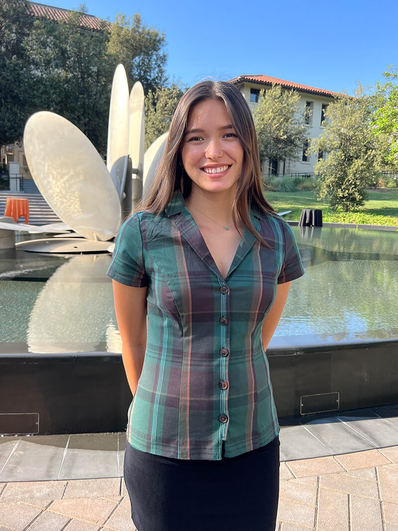 Sophia is wearing a green plaid shirt and is standing in front of a fountain at the Oxy Campus