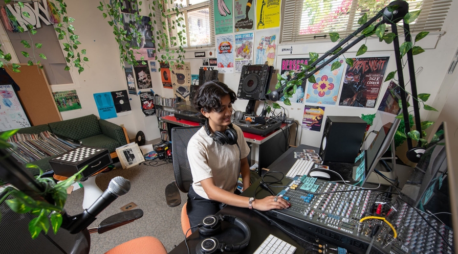 An Occidental College student in the KOXY radio station, at the DJ booth with plants all around