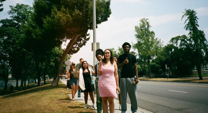 students standing on a sidewalk by a road, giving thumbs up signs