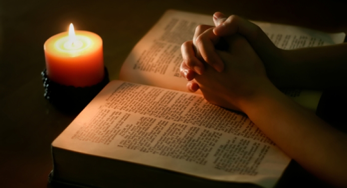 Two hands folded over a Bible illuminated by candlelight