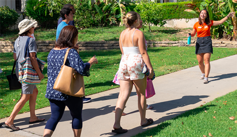 Student tour guide in orange T shirt leads visitors through green spaces at Oxy along a sidewalk