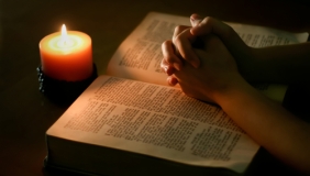 Two hands folded over a Bible illuminated by candlelight