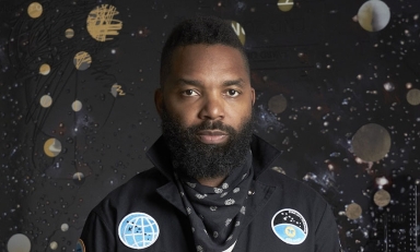 Tavares Strachan in a black suit with cosmic imagery behind him on a black background