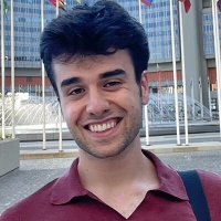 Peter is wearing a red shirt and is standing in front of a fountain and several flags